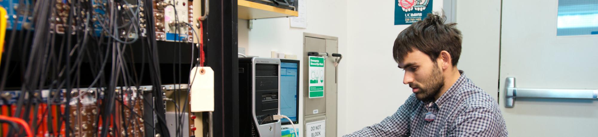 A UC Davis researcher, who is Hispanic and a man, works at computer and is seated next to several servers with cables hanging from them.